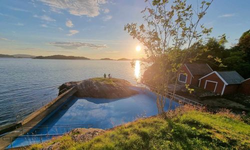 Bildet viser et idyllisk kystlandskap ved solnedgang, med blikkstille sjø og varme solstråler som speiler seg i vannflaten. I forgrunnen ser vi et utendørs badebasseng, bygd inn i terrenget og omkranset av rekkverk. Til høyre står et tre med nyutsprungne blader, og bak det ser vi noen røde bygninger med hvite karmer – sannsynligvis eldre sjøboder eller småhus knyttet til stedet. Midt i bildet står tre personer på en svaberg-tunge som stikker ut i sjøen. Utsikten strekker seg utover øyer og holmer i det fjerne. Bildet er tatt fra husmannsplassen Træ ved Follese på Askøy.